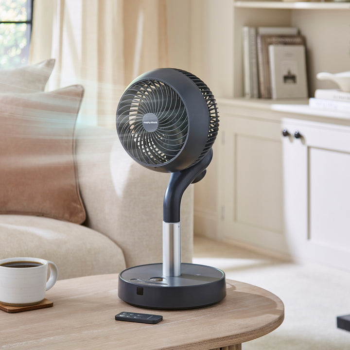 Black stand fan on a wooden table in a living room setting with a cup of coffee and remote control.