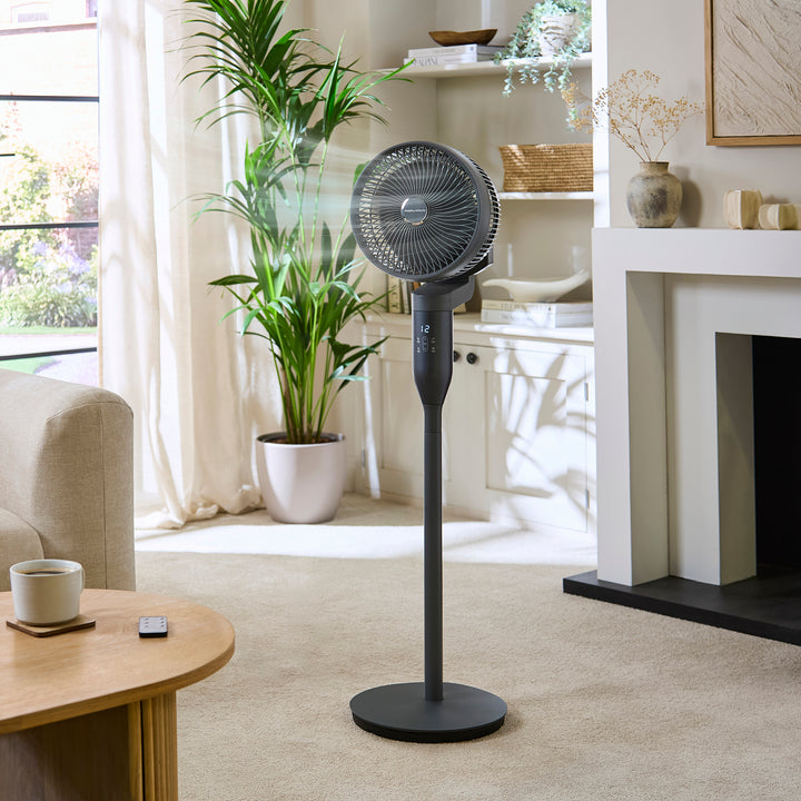 Black stand fan in a living room setting with a plant and table.