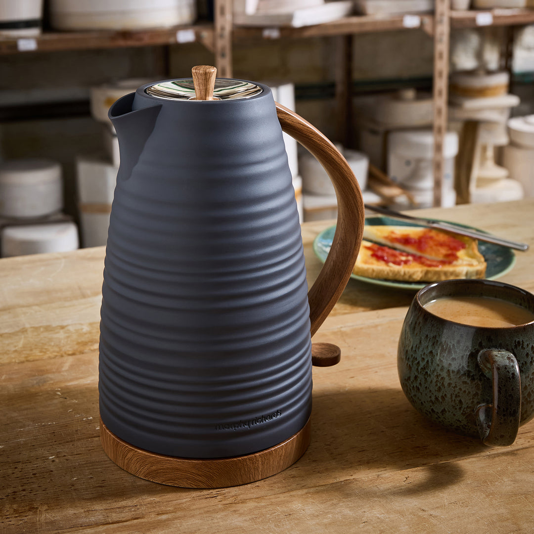 Blue ceramic Kettle with wooden handle on a wooden table with a cup of coffee and plate of food in a workshop setting.