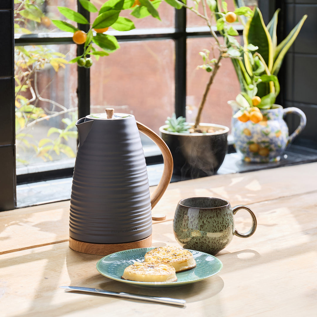 Gray ceramic kettle, green mug, and plate with crumpets on a wooden table with plants in the background.