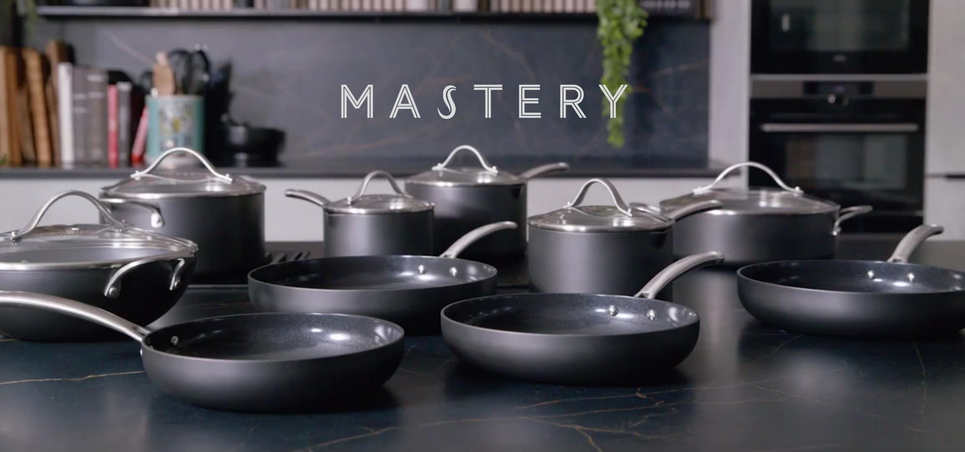 A collection of dark gray non-stick cookware, including pans and pots with glass lids, arranged on a dark countertop in a modern kitchen setting. The word 'MASTERY' is displayed in white text across the center.