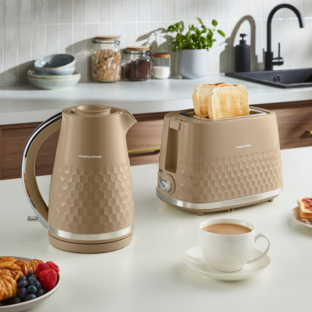 A beige kettle and matching toaster set on a white kitchen countertop, with toast in the toaster, a cup of coffee, and breakfast items in a real kitchen setting.