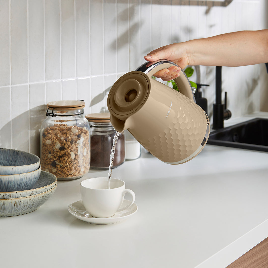A beige electric kettle with a textured honeycomb pattern is being used to pour hot water from a white teacup on a saucer in a bright, modern kitchen.