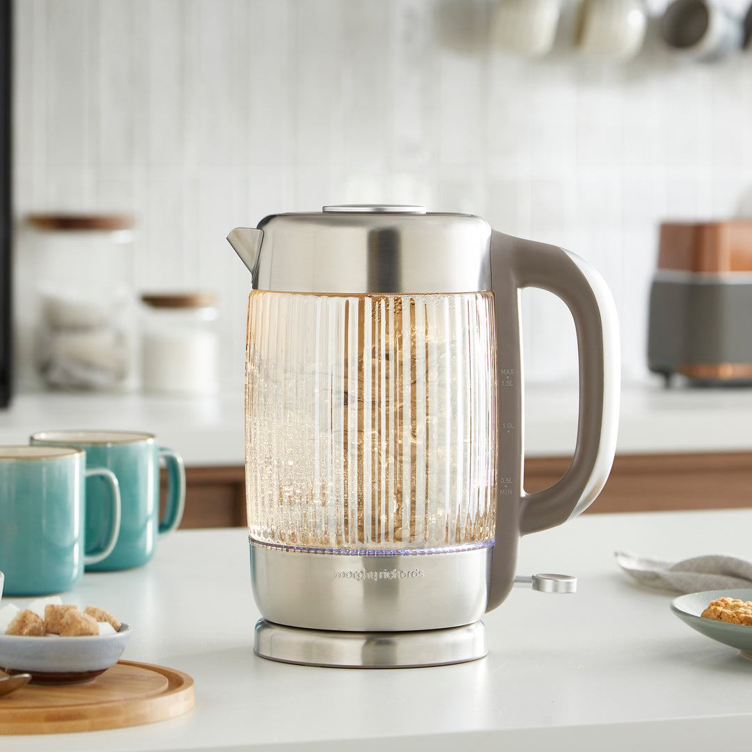Clear glass electric kettle on a kitchen counter with mugs and kitchen items in the background