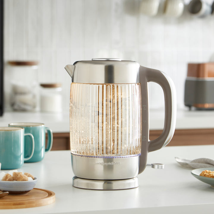 Clear glass electric kettle on a kitchen counter with mugs and kitchen items in the background