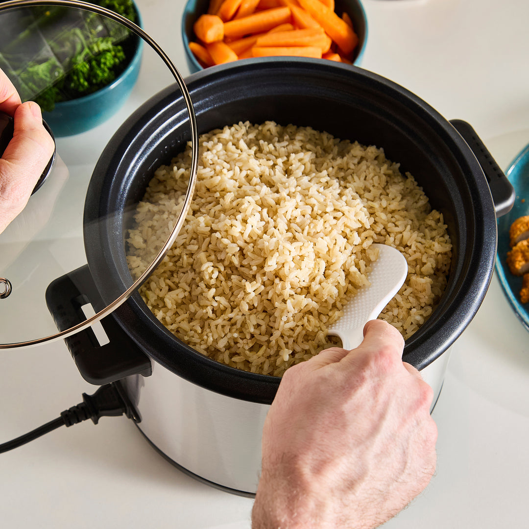 Person using a rice cooker with cooked rice inside, surrounded by vegetables on a table.