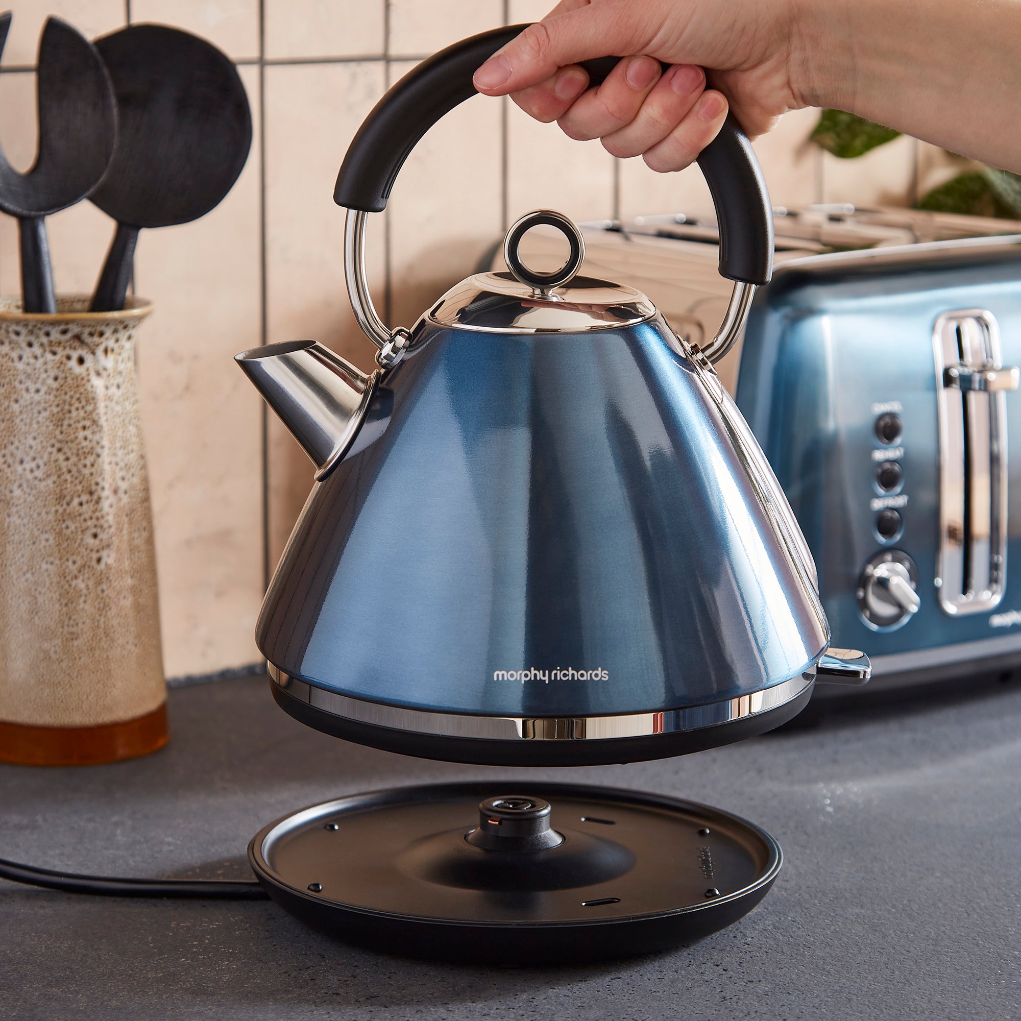 Blue kettle being held by a hand on a kitchen counter with a toaster and utensils in the background.