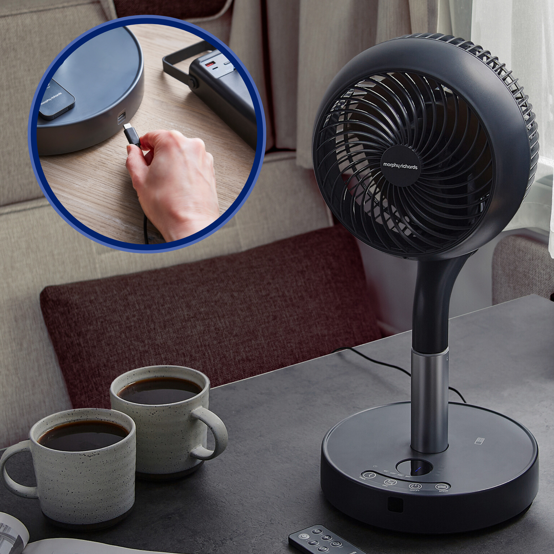 Black handheld fan on a table with two cups of coffee and a remote control.