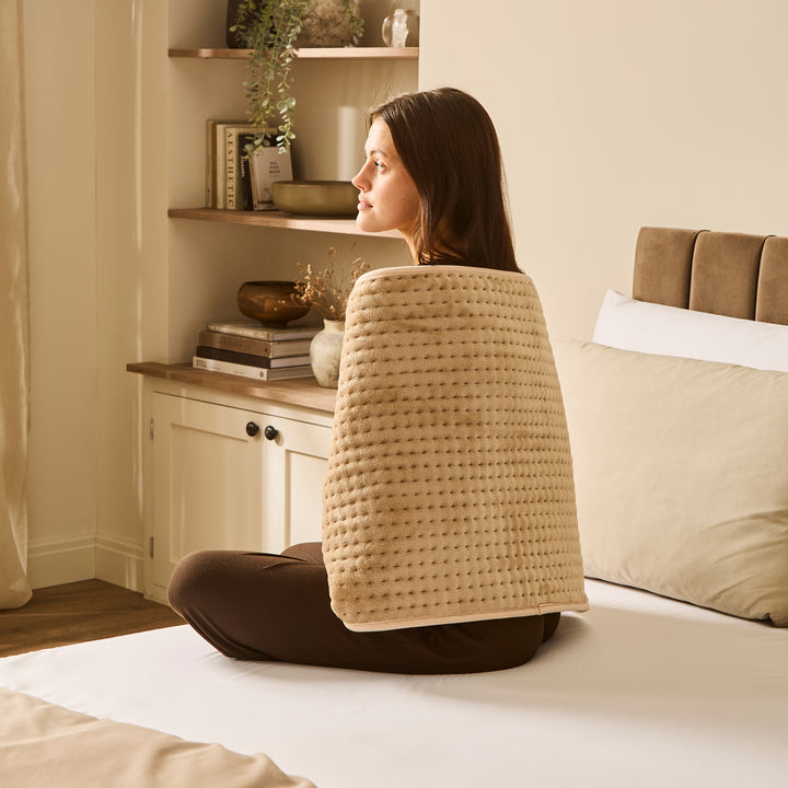 Woman sitting on a bed with a heat pad draped over shoulder and back