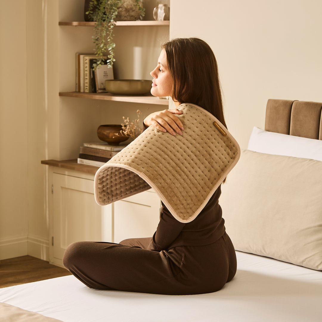 Woman sitting on a bed holding a beige heat pad on shoulder. 