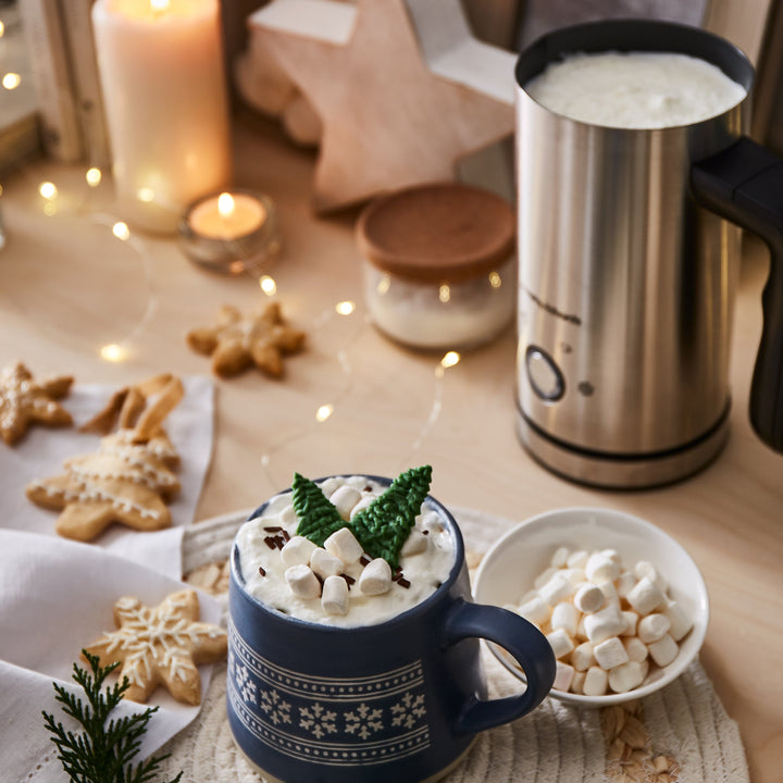 Hot chocolate with marshmallows in a blue mug on a festive table with cookies and a milk frother