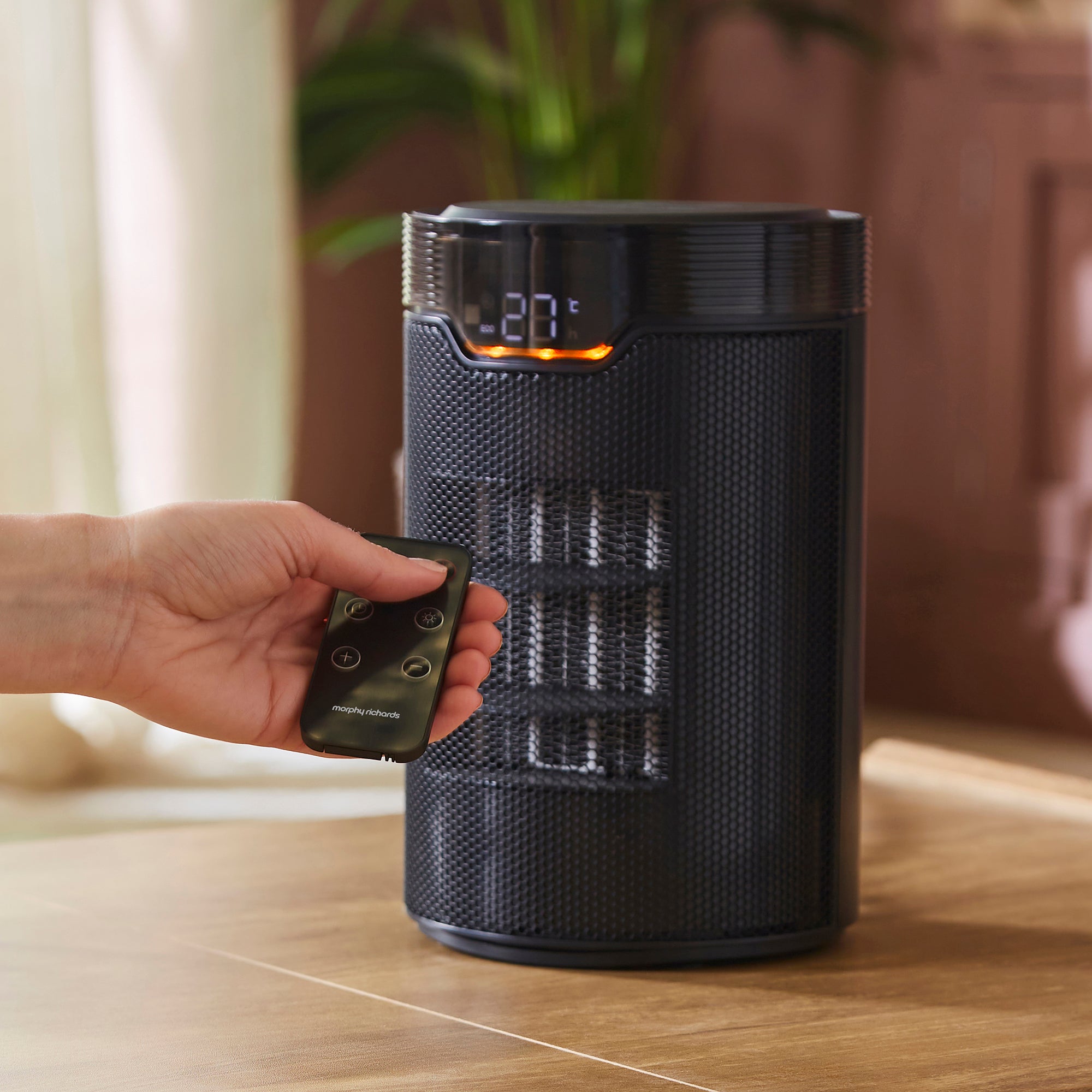 Black fan heater with digital display and remote control held by a hand in a home setting.