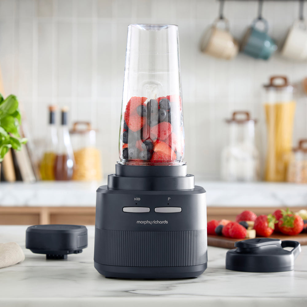 A Morphy Richards blender with a clear jar filled with strawberries and blueberries, set on a marble countertop in a kitchen. The black blender base is shown with its lid off on the left and the detached lid on the right.