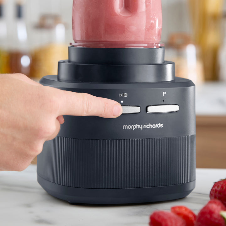 A close-up of a hand pressing the power button on a Morphy Richards blender base, with a pink smoothie in the jar and strawberries on a marble countertop in a kitchen setting.