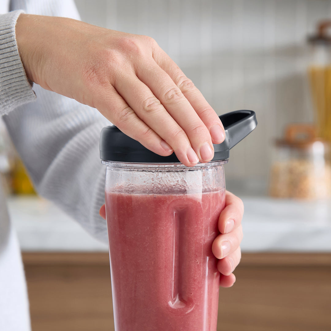 A close-up of a person's hands holding a clear plastic smoothie cup with a black lid, filled with a pink smoothie, in a bright kitchen setting.