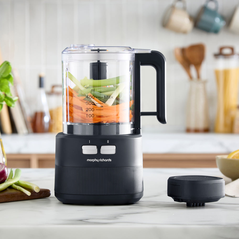 A Morphy Richards countertop food processor with a clear jar containing carrots and celery, shown on a light marble kitchen counter with the lid placed beside it.