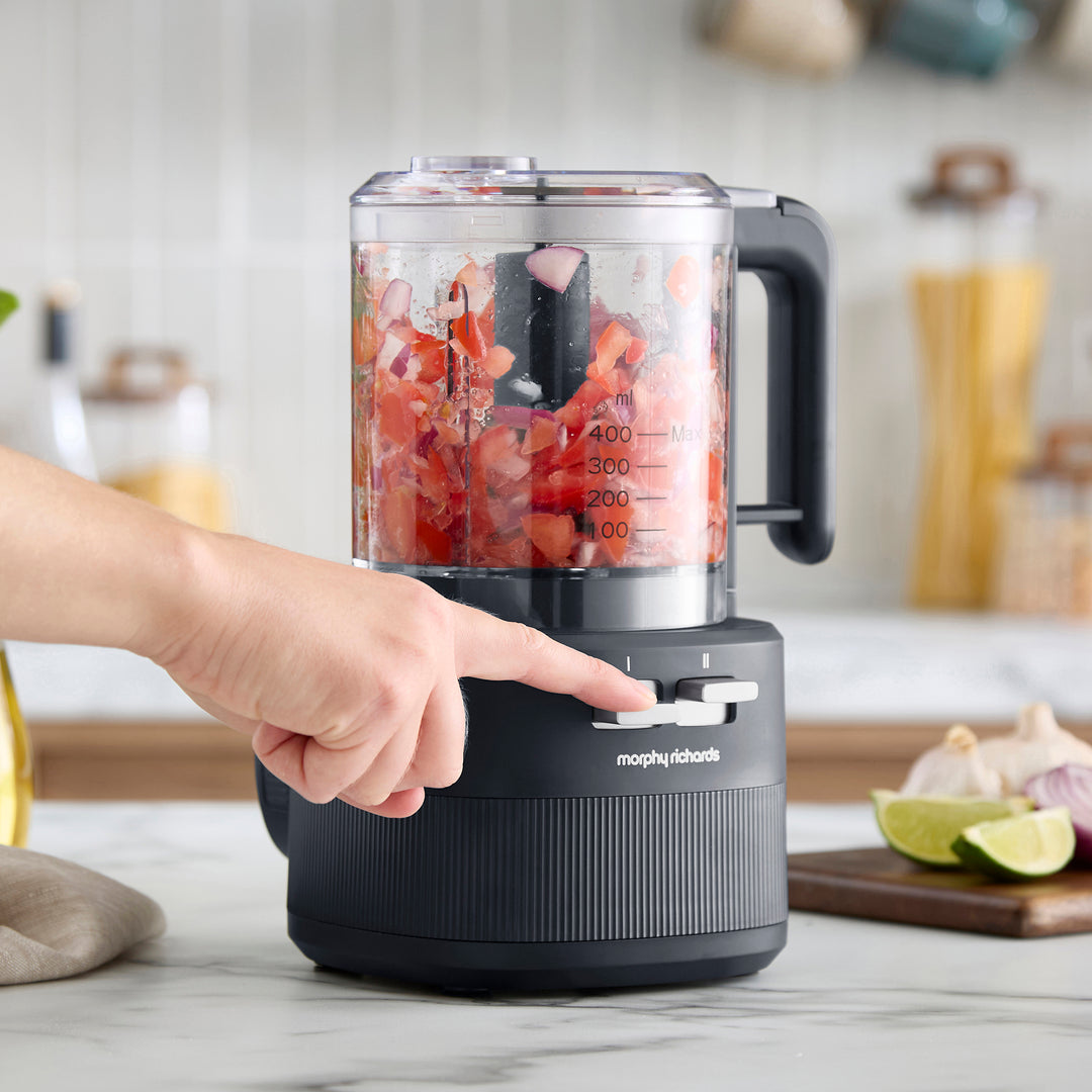 A Morphy Richards food processor with a clear jar filled with chopped red vegetables, being operated by a person whose hand and arm are visible, in a bright kitchen setting.