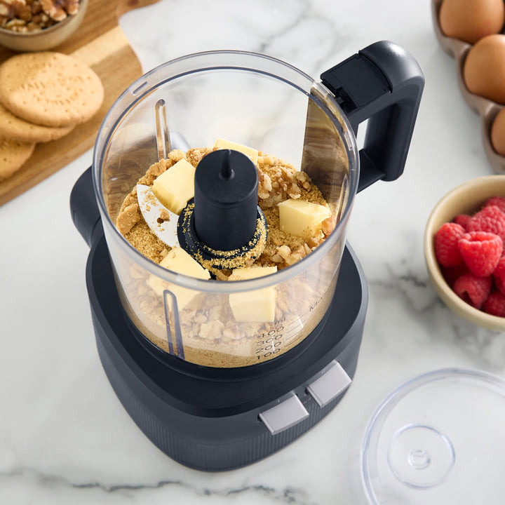 A countertop food processor with a clear bowl and dark grey base, shown with butter and chopped nuts inside. The scene is styled on a white marble surface with baking ingredients and utensils around it.