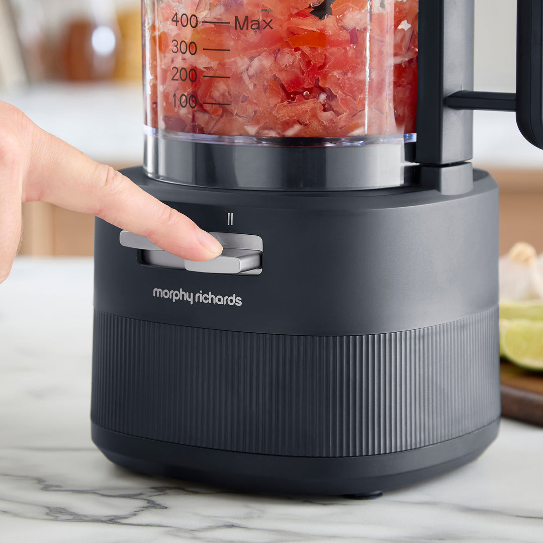 A close-up of a Morphy Richards foods processor with a dark grey base and a clear pitcher filled with chopped red tomatoes. A person's finger is pressing the start button on the front.
