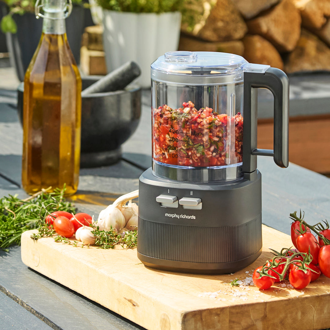 A countertop food processor filled with chopped red vegetables, placed on a wooden cutting board outdoors with tomatoes, garlic, herbs, and a bottle of oil nearby. The Morphy Richards brand name is visible on the base.