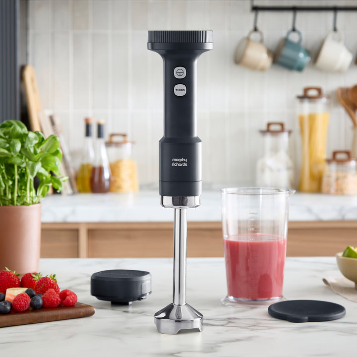 A black and silver Morphy Richards immersion blender stands on a white marble kitchen countertop, with a glass of smoothie and berries.