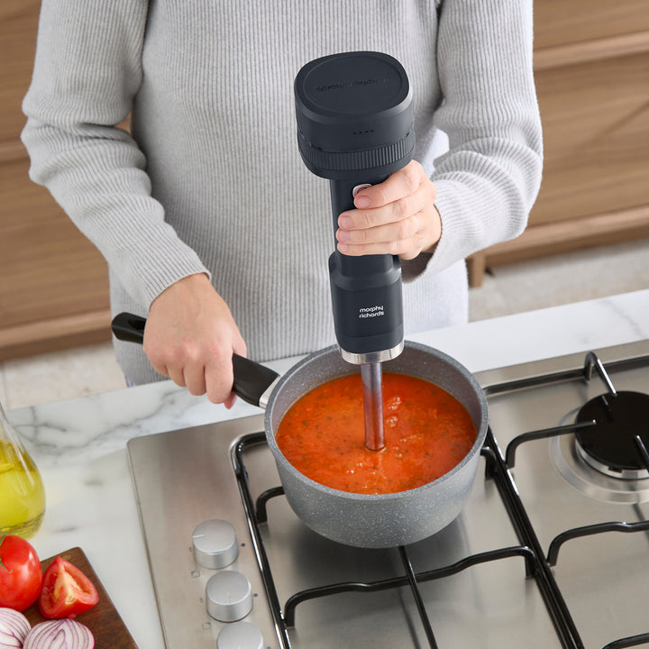 A person in a grey sweater uses a black immersion blender to mix tomato soup in a grey pot on a stovetop in a kitchen setting.