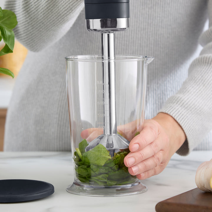 A person using an immersion blender to puree green leafy vegetables in a clear plastic blending cup. 