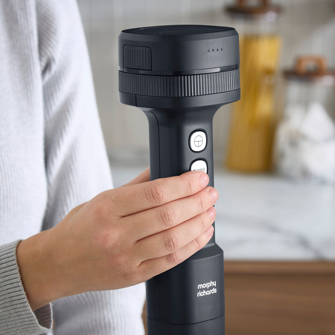 A close-up of a person holding a hand blender with a hand over one of its buttons, set against a blurred kitchen background.