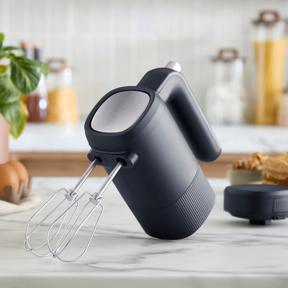 A modern hand-held electric mixer with a matte dark grey body and a stainless steel wire whisk attachment, shown against a solid white background.