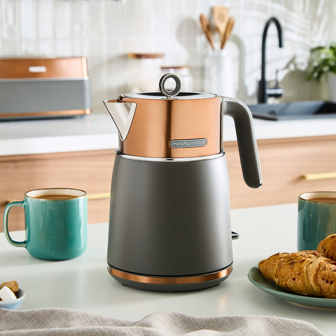 Grey and copper jug kettle on a kitchen counter with mugs and pastries.