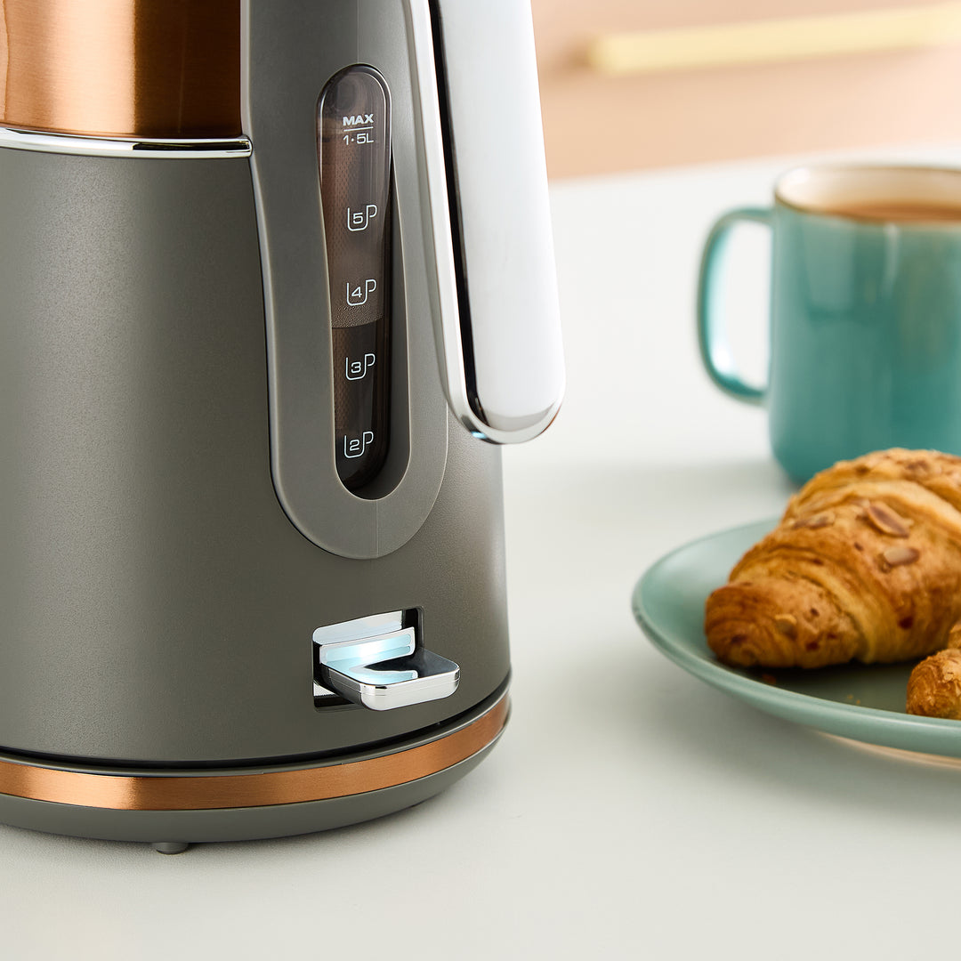 Grey electric kettle with copper accents on a kitchen counter with a croissant and mug.