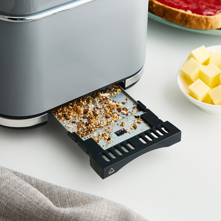 Grey toaster with open crumb tray on a kitchen counter with bread and butter in the background.