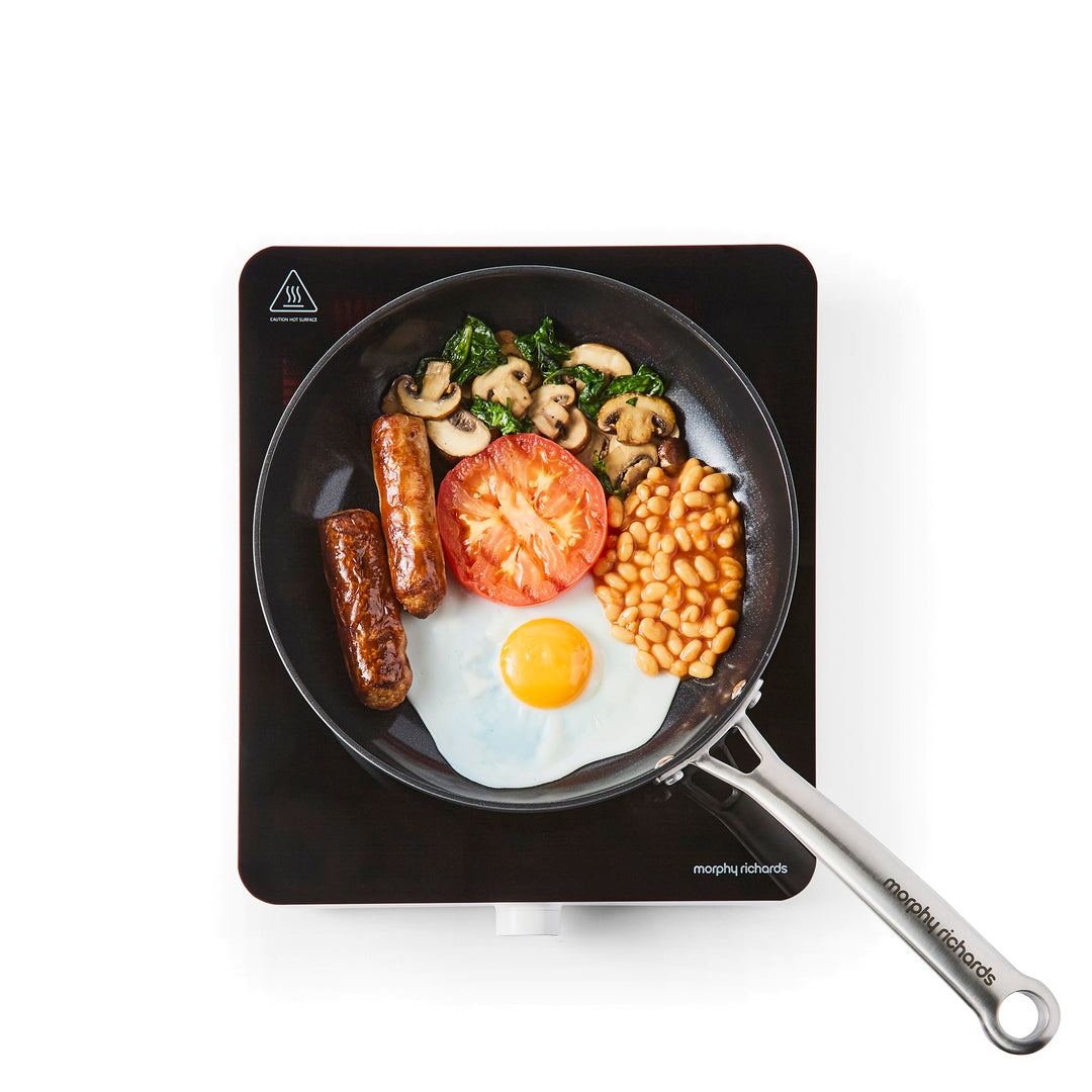 single induction hob with frying pan on top filled with full english breakfast ingredients on white background