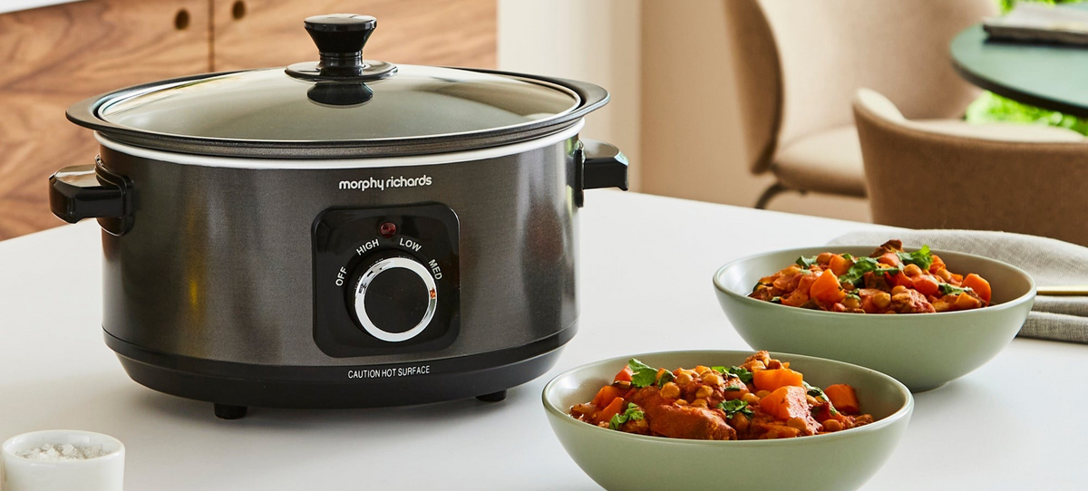 A Morphy Richards slow cooker in dark gray sits on a white table, with two bowls of cooked stew in the foreground and a softly blurred dining room background.