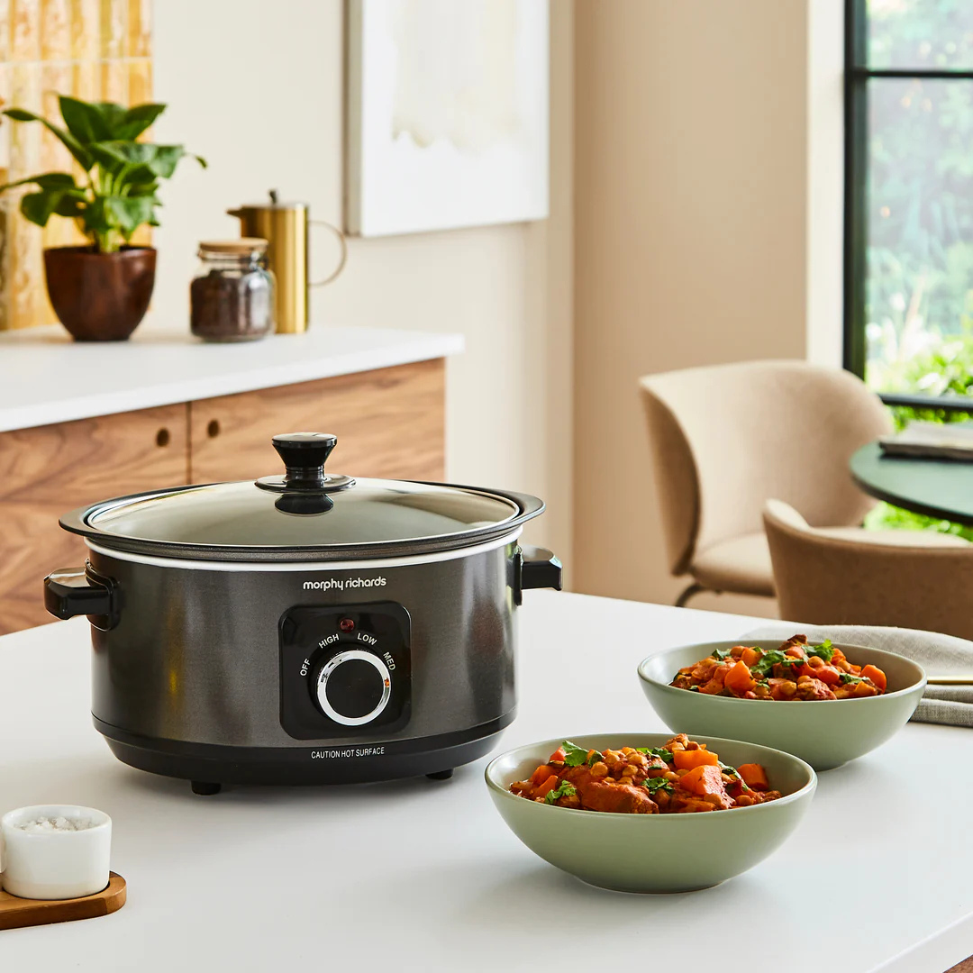 A slow cooker pot warmer sits on a white table in a bright kitchen setting, with two bowls of prepared food in front and a countertop with plants and decor in the background.